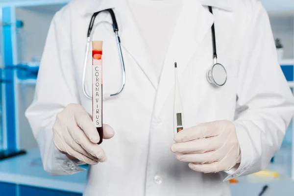 Vista recortada del científico en guantes de látex con termómetro digital y tubo de ensayo con letras de coronavirus - foto de stock