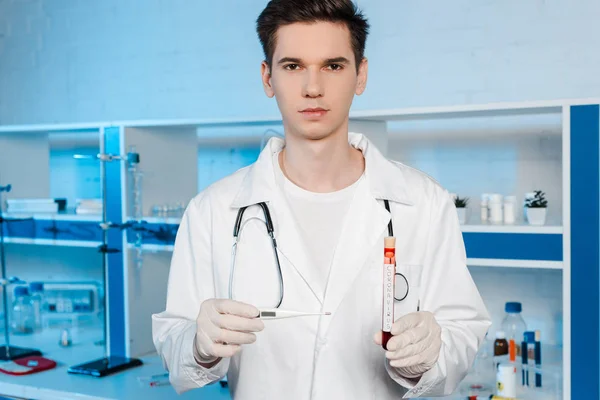 Bellissimo scienziato in guanti di lattice con termometro digitale e provetta con scritta coronavirus — Foto stock