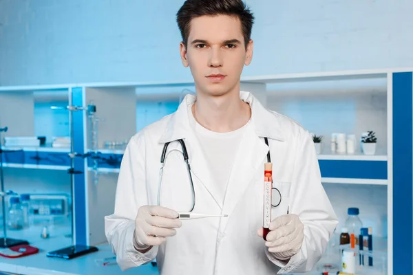 Handsome scientist in latex gloves holding digital thermometer and test tube with coronavirus lettering — Stock Photo
