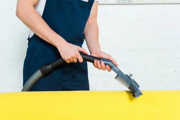 Vue recadrée du nettoyeur en salopette enlever la poussière sur le canapé avec aspirateur — Photo de stock