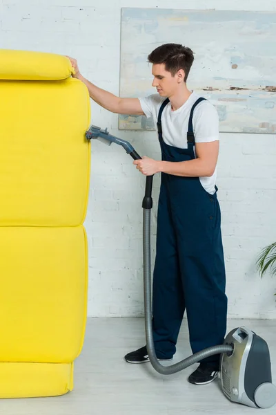 Vue latérale du nettoyeur en uniforme nettoyage à sec canapé moderne avec aspirateur — Photo de stock