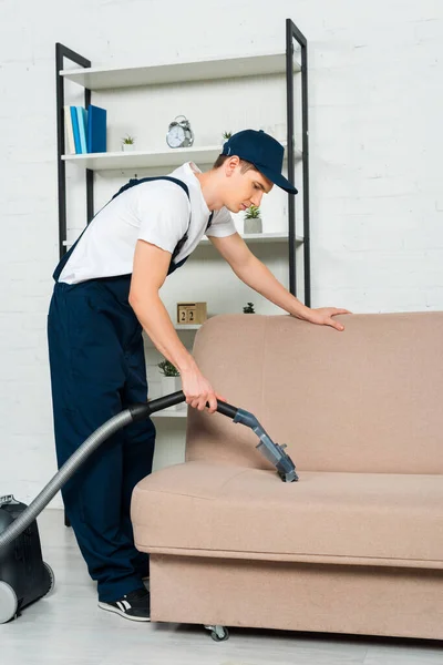 Side view of cleaner in cap removing dust on sofa with vacuum cleaner — Stock Photo