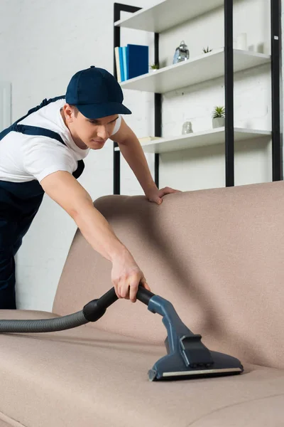 Young cleaner in cap removing dust on sofa with vacuum cleaner — Stock Photo