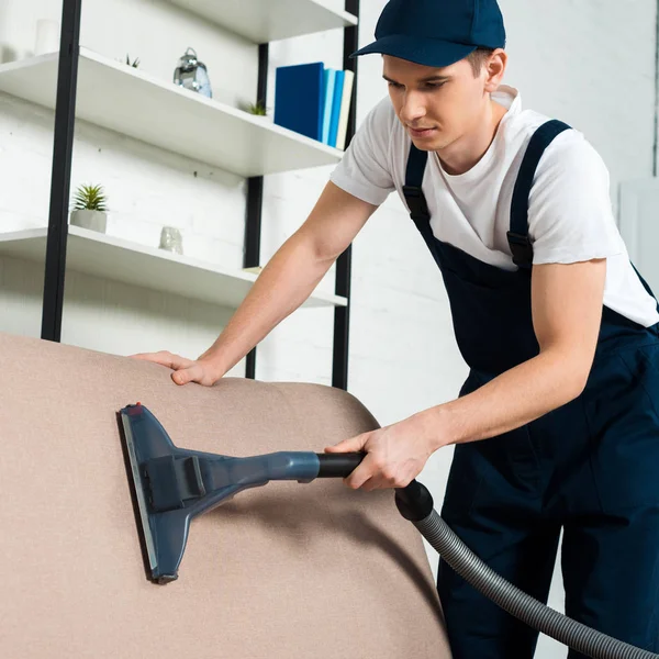 Young handsome cleaner in cap removing dust on sofa with vacuum cleaner — Stock Photo