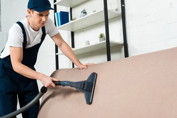 Junge Reinigungskraft in Kappe und Uniform Staubsaugen auf dem Sofa mit Staubsauger — Stockfoto