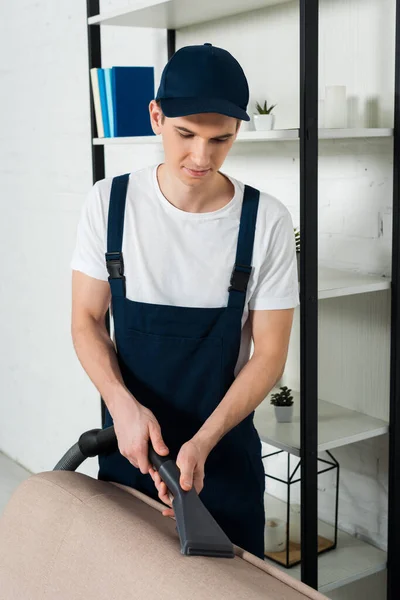 Jeune homme en chapeau et uniforme enlever la poussière sur le canapé avec aspirateur — Photo de stock