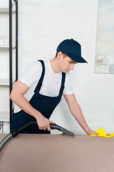 Young man in cap and overalls removing dust on sofa with vacuum cleaner and cleaning cloth — Stock Photo