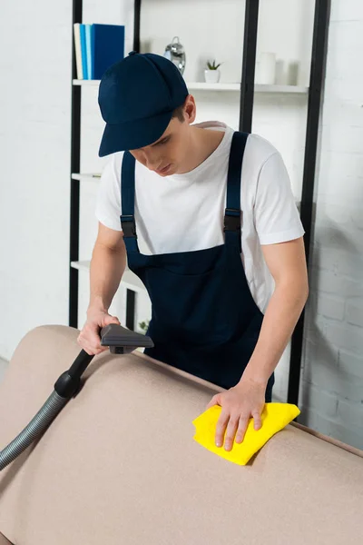 Hombre en gorra y mono sofá de limpieza con aspiradora y paño de limpieza - foto de stock