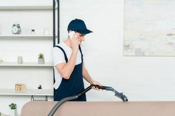 Cleaner in cap and overalls cleaning sofa with vacuum cleaner and talking on smartphone — Stock Photo