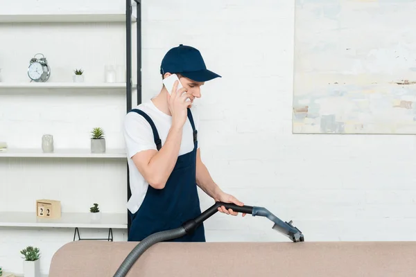 Limpiador en gorra y mono limpieza sofá con aspiradora y hablando en el teléfono inteligente - foto de stock