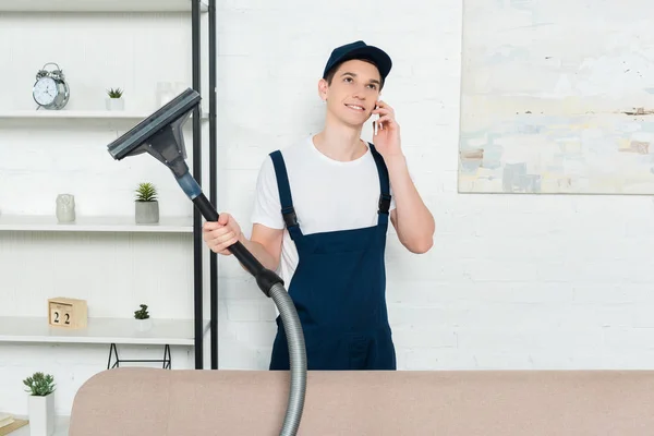 Happy cleaner in cap and overalls holding vacuum cleaner and talking on smartphone — Stock Photo