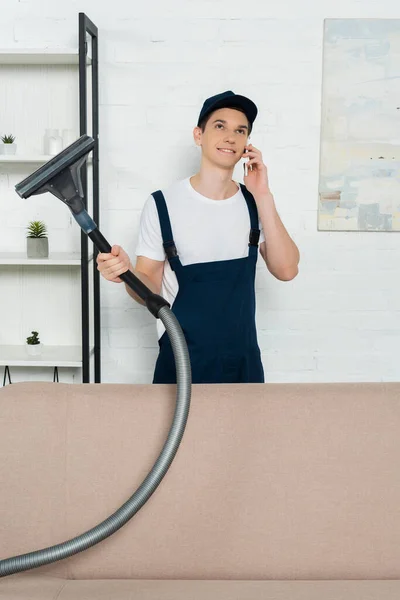 Cheerful cleaner in cap and overalls holding vacuum cleaner and talking on smartphone — Stock Photo