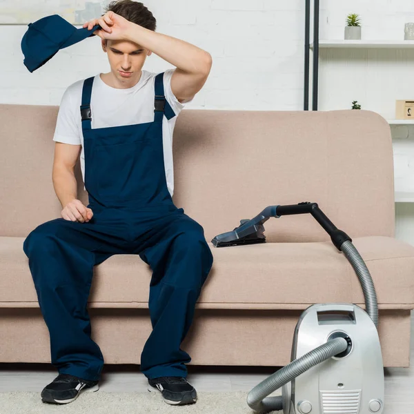 Tired cleaner in overalls holding cap while sitting on sofa near vacuum cleaner — Stock Photo