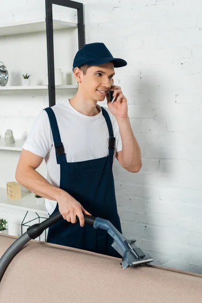 Jeune nettoyeur souriant en bonnet et salopette tenant aspirateur et parlant sur smartphone — Photo de stock