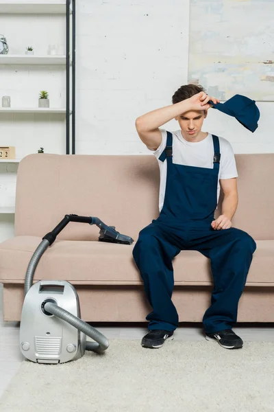 Exhausted cleaner in overalls holding cap while sitting on sofa near vacuum cleaner — Stock Photo