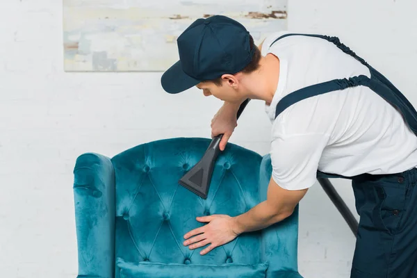 Junge Reinigungskraft in Uniform und Mützenreinigung moderner Sessel mit Staubsauger — Stockfoto