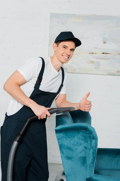 Heureux nettoyeur dans le chapeau montrant pouce vers le haut tout en nettoyant à sec fauteuil bleu avec aspirateur — Photo de stock