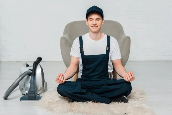 Happy cleaner in cap meditating on carpet near vacuum cleaner and armchair — Stock Photo