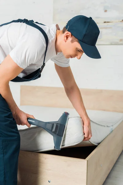 Young cleaner in cap removing dust on mattress with vacuum cleaner — Stock Photo