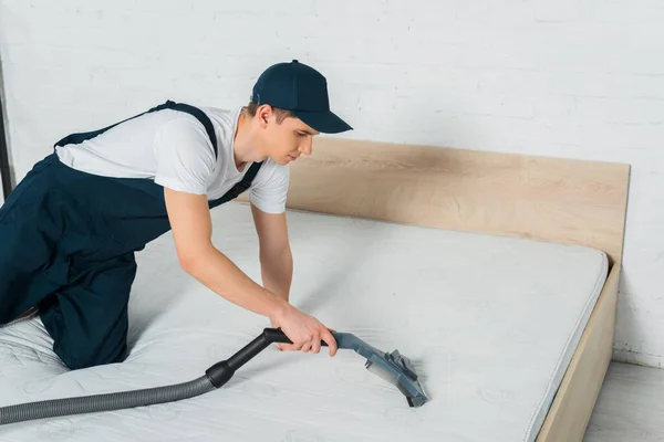 Handsome cleaner in cap removing dust on mattress with vacuum cleaner — Stock Photo