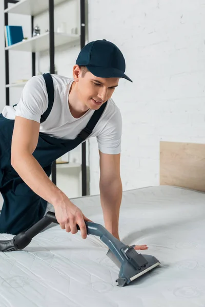 Happy cleaner in cap removing dust on mattress with vacuum cleaner — Stock Photo