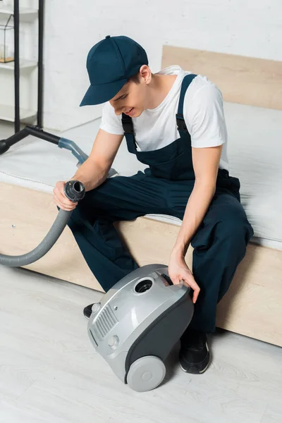 Happy cleaner in cap holding hose near vacuum cleaner in bedroom — Stock Photo
