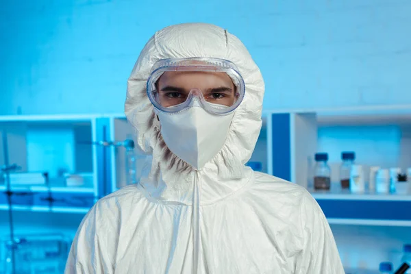 Científico en traje de materiales peligrosos, máscara médica y gafas mirando a la cámara - foto de stock