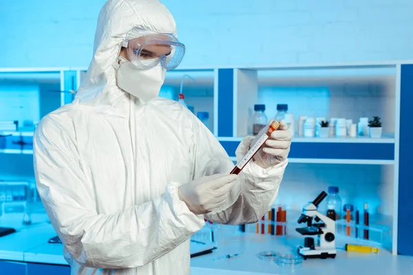 Científico en traje de materiales peligrosos, guantes de látex y gafas con tubo de ensayo con letras de coronavirus - foto de stock