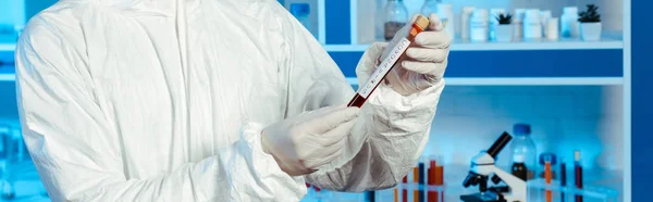 Panoramic shot of scientist in hazmat suit and latex gloves holding test tube with coronavirus lettering — Stock Photo