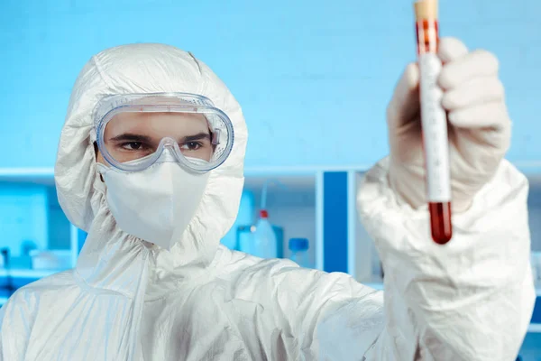 Enfoque selectivo del científico en traje de materiales peligrosos y gafas que sostienen el tubo de ensayo con letras de coronavirus - foto de stock