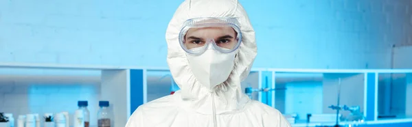 Панорамний знімок вченого в косметичному костюмі, медична маска та окуляри, що дивляться на камеру — стокове фото