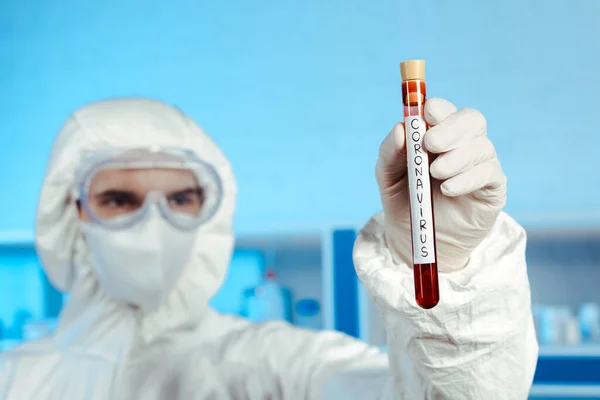 Selective focus of scientist in hazmat suit, medical mask and goggles holding test tube with coronavirus lettering — Stock Photo