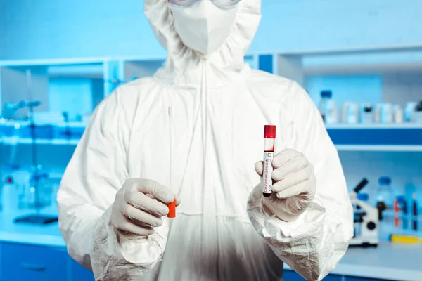 Vue recadrée du scientifique en gants de latex et masque médian tenant une éprouvette avec lettrage au coronavirus — Photo de stock