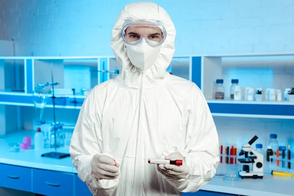 Científico en traje de materiales peligrosos, máscara médica y gafas que sostiene la muestra con letras de coronavirus - foto de stock
