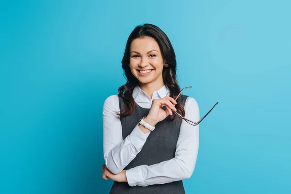 Alegre mujer de negocios sosteniendo gafas mientras sonríe a la cámara sobre fondo azul - foto de stock