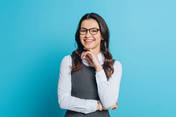 Cheerful businesswoman smiling at camera while holding hand near chin on blue background — Stock Photo