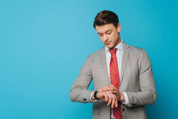 Guapo y serio hombre de negocios comprobando el tiempo en el reloj de pulsera sobre fondo azul - foto de stock