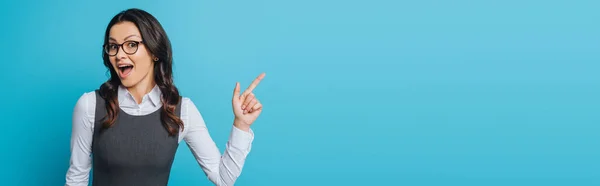 Panoramic shot of amazed businesswoman in eyeglasses pointing with finger isolated on blue — Stock Photo