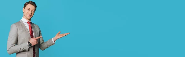 Панорамний знімок усміхненого бізнесмена, який вказує пальцем, стоячи з відкритою рукою і дивлячись на камеру ізольовано на синьому — стокове фото