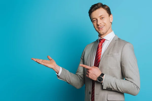 Sonriente hombre de negocios señalando con el dedo mientras está de pie con el brazo abierto y mirando a la cámara en el fondo azul - foto de stock