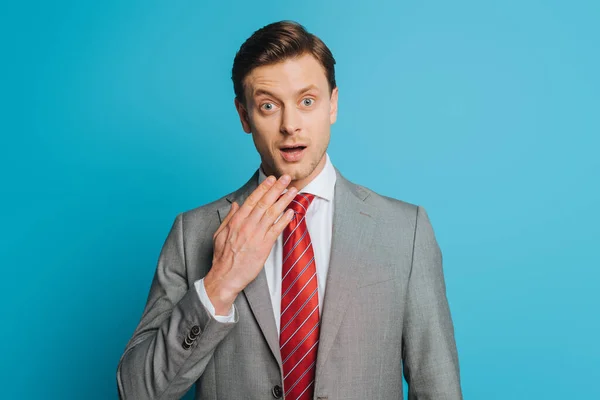 Shocked businessman touching chin while looking at camera with wide open eyes on blue background — Stock Photo