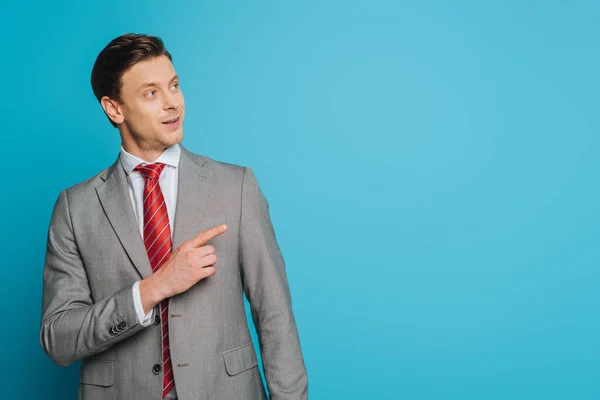 Smiling businessman looking away and pointing with finger on blue background — Stock Photo