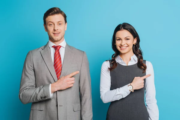 Alegre hombre de negocios y mujer de negocios señalando con los dedos mientras sonríe a la cámara sobre fondo azul - foto de stock