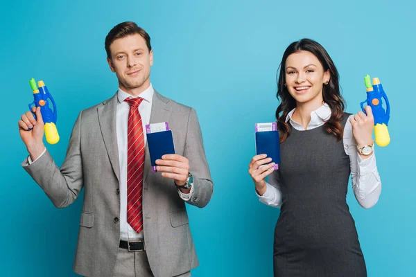 Two excited businesspeople holding passports, air tickets and water guns on blue background — Stock Photo