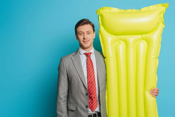 Happy businessman holding inflatable mattress while looking at camera on blue background — Stock Photo