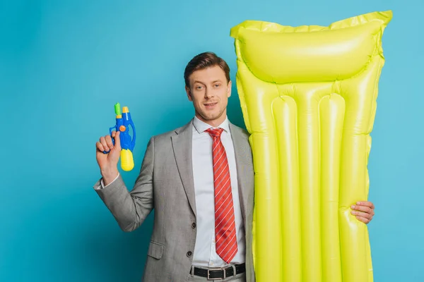 Sonriente hombre de negocios sosteniendo colchón inflable y pistola de agua sobre fondo azul - foto de stock