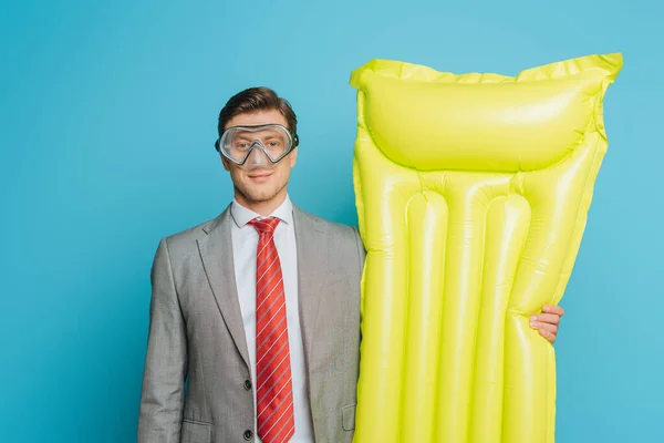Sonriente hombre de negocios en máscara de buceo sosteniendo colchón inflable sobre fondo azul - foto de stock