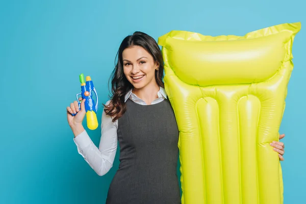 Sonriente mujer de negocios sosteniendo colchón inflable y pistola de agua sobre fondo azul - foto de stock