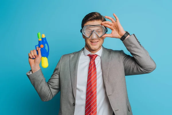 Alegre hombre de negocios en la máscara de buceo sosteniendo pistola de agua sobre fondo azul - foto de stock