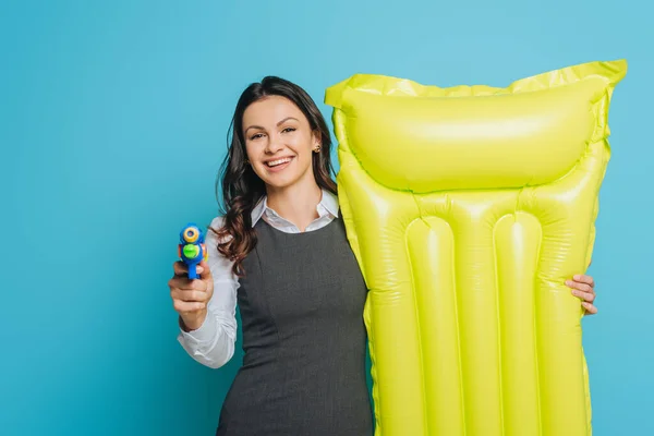 Fröhliche Geschäftsfrau hält aufblasbare Matratze in der Hand und zeigt mit Wasserpistole auf Kamera auf blauem Hintergrund — Stockfoto